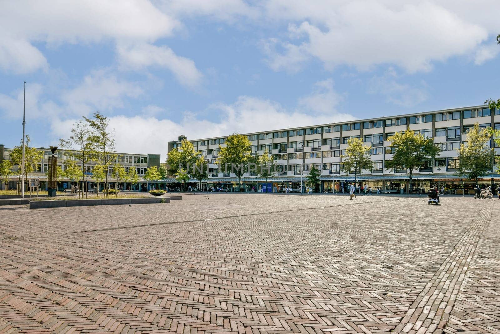 A spacious urban plaza featuring modern buildings and lush greenery, bustling with people and surrounded by clear blue skies, showcasing contemporary city life.