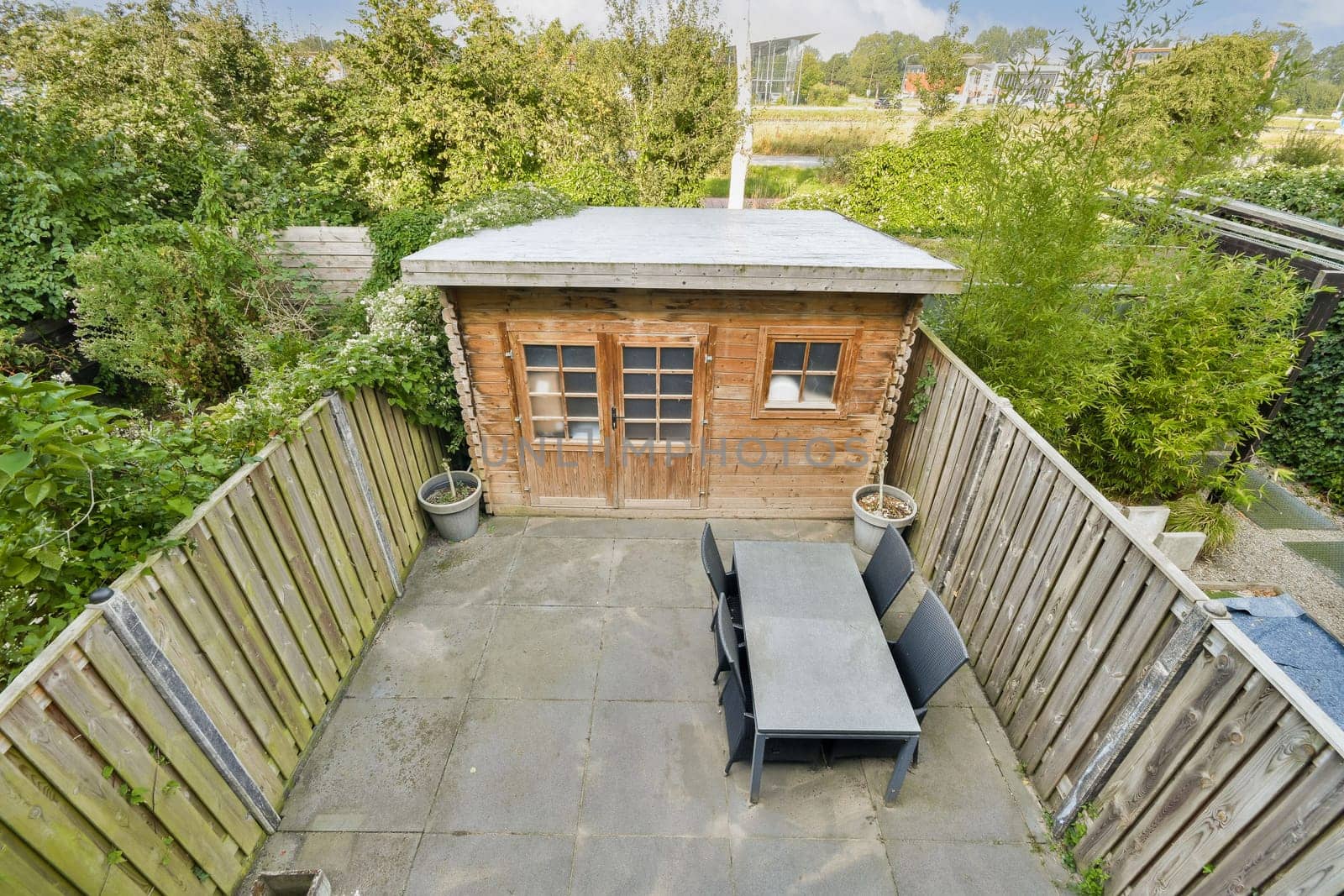 A stylish wooden garden shed surrounded by greenery, featuring a patio area with a table and chairs, perfect for outdoor relaxation and storage.