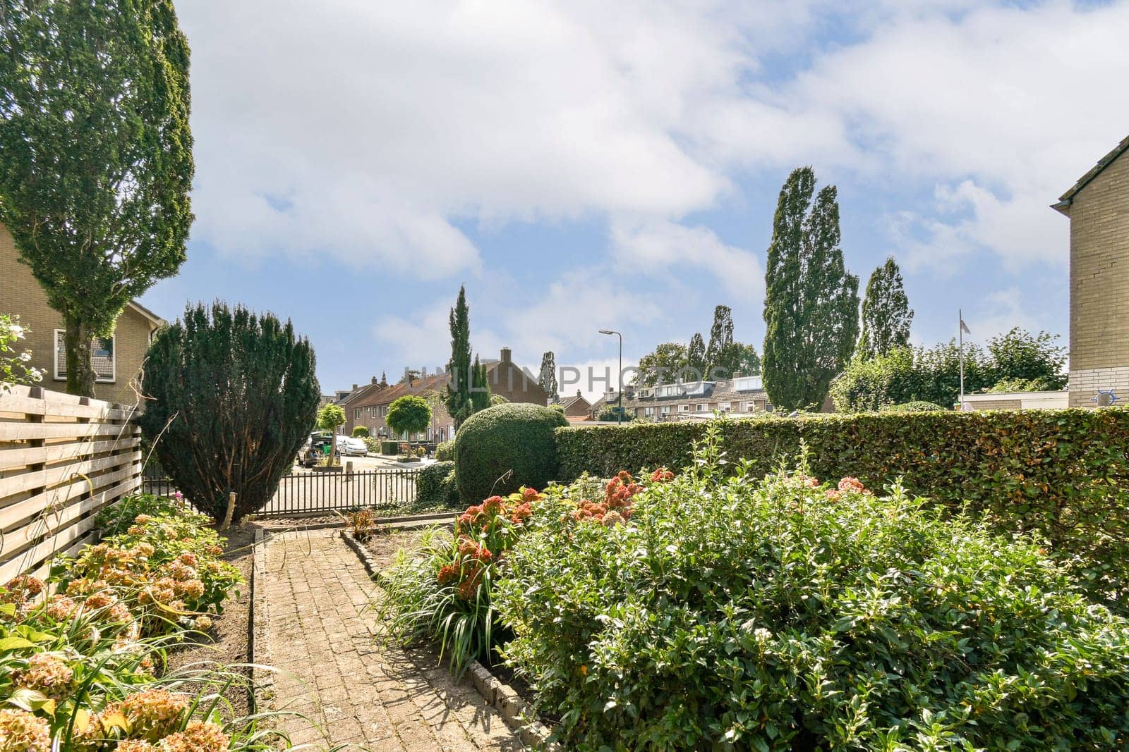 A serene garden pathway is framed by lush greenery and vibrant flowers, leading through a peaceful residential area under a clear blue sky.