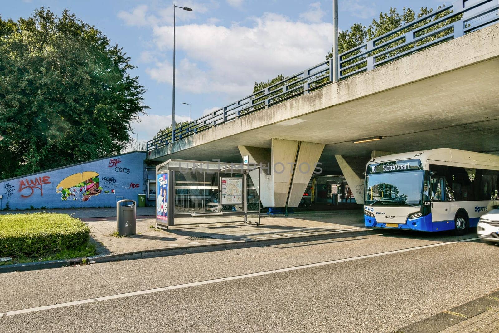 A modern bus stop in an urban environment with vibrant street art on the wall, surrounded by greenery and a busy roadway.