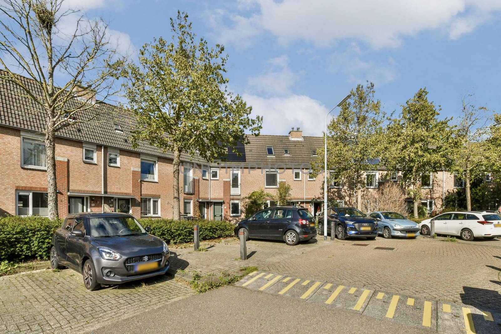A view of a residential parking area featuring multiple parked cars surrounded by grassy patches and trees under a clear blue sky.
