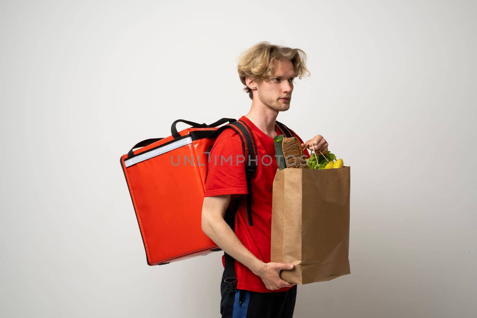 Side view handsome Caucasian grocery delivery courier man in red uniform with grocery bag with fresh fruit and vegetable and frozen food carrier bag on a shoulder
