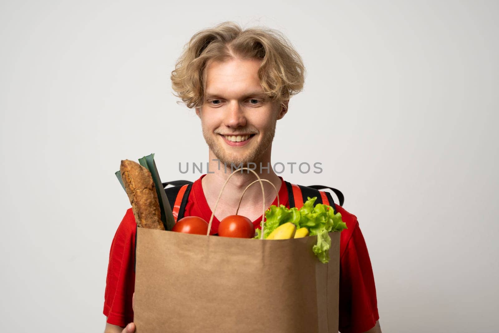 Delivery. Handsome Caucasian grocery delivery courier man in red uniform with grocery bag with fresh fruit and vegetable and frozen food carrier bag on a shoulder