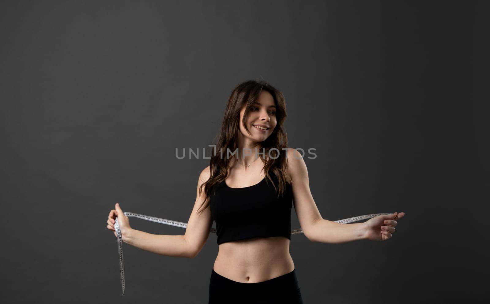 Beautiful brunette young woman in black outfit with measuring tape. Measuring tape on the waist. Beautiful fitness woman is in the studio