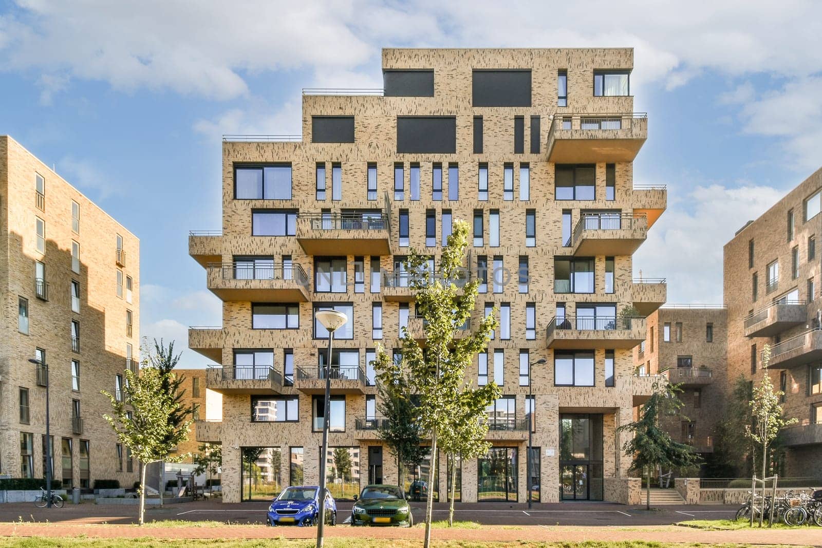 A uniquely designed modern apartment building features a textured facade and multiple balconies, set against a bright sky with trees and vehicles in view.
