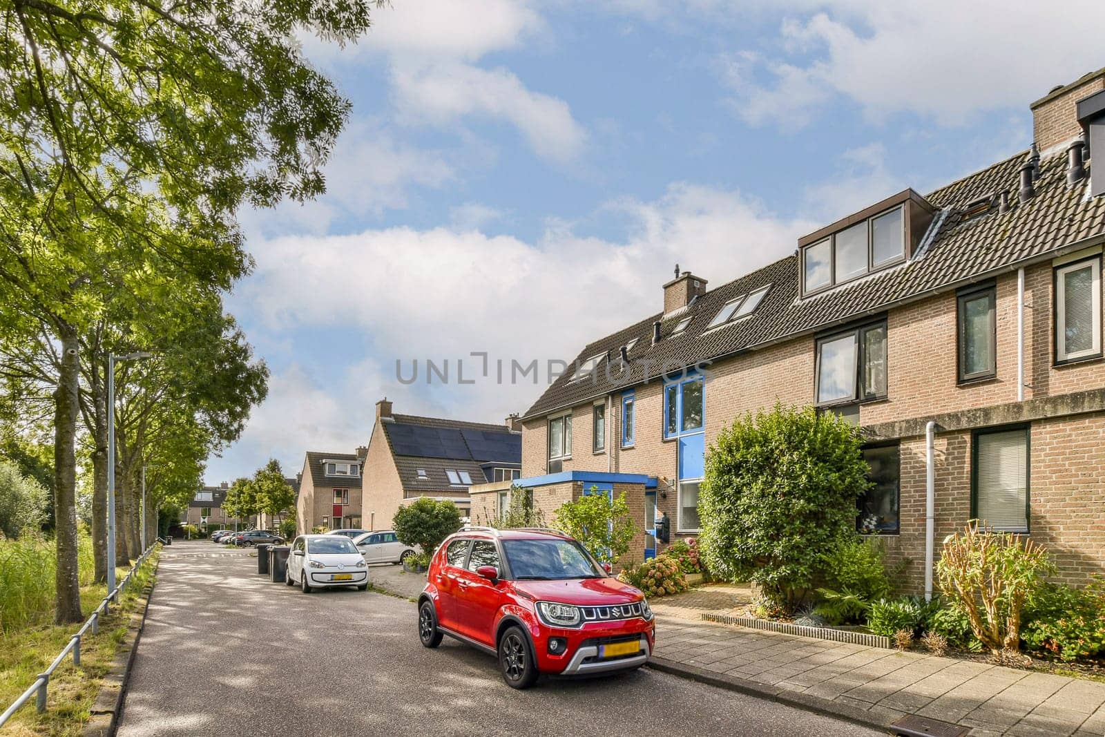 A scenic view of a residential area featuring modern buildings, parked cars, and green trees lining the street. The vibrant red car stands out against the backdrop.