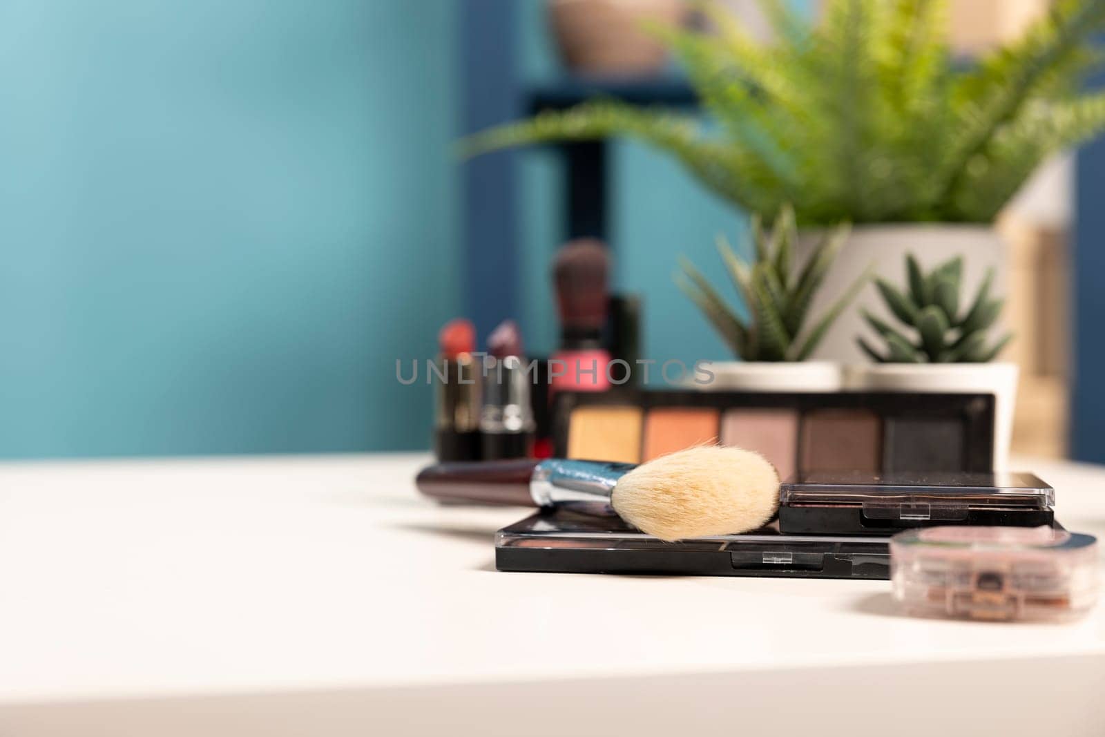 Selective focus on a brush placed on a white table along with other cosmetic products. A well organized beauty studio setup featuring a neatly arranged desk with makeup, brushes, and mini plants.