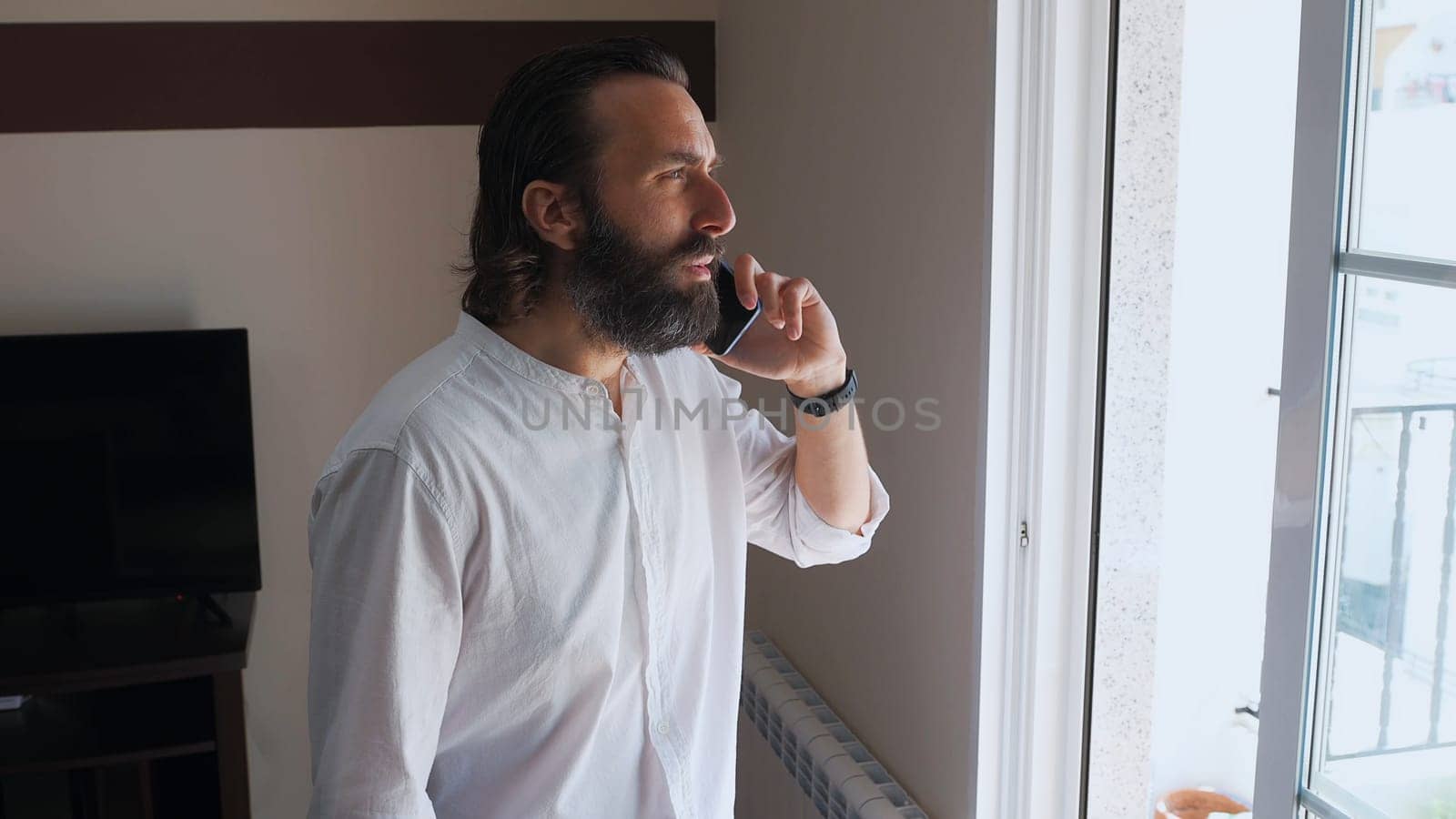 Young man standing beside a window, speaking on his smartphone with a focused expression. Wearing a white shirt, he appears businesslike in his home office setting.