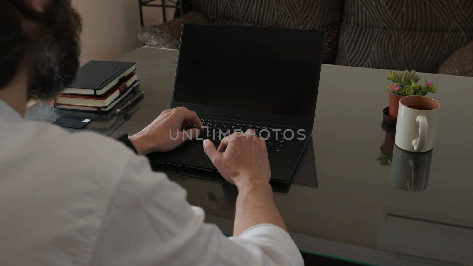 A man in a professional workspace uses a laptop computer while surrounded by notebooks, a plant, and a coffee mug. The scene suggests focus, productivity, and a comfortable office environment.