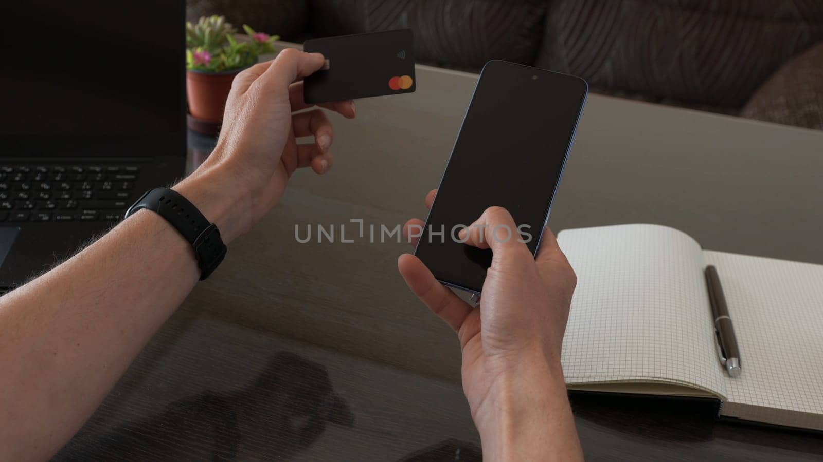A person operates a smartphone alongside a credit card, surrounded by workspace essentials, emphasizing modern technology and business convenience.
