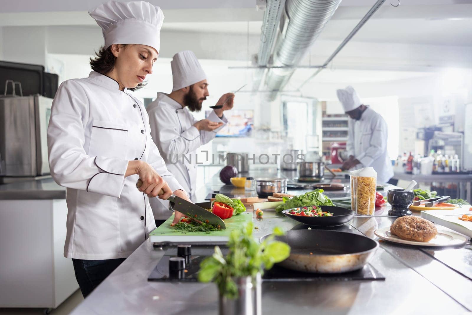 Focused female chef cutting small pieces of flavory herbs for a dish on the stove, mincing rosemary or basil on the cutting board. Woman cook using greenery and condiments for tasty meal.
