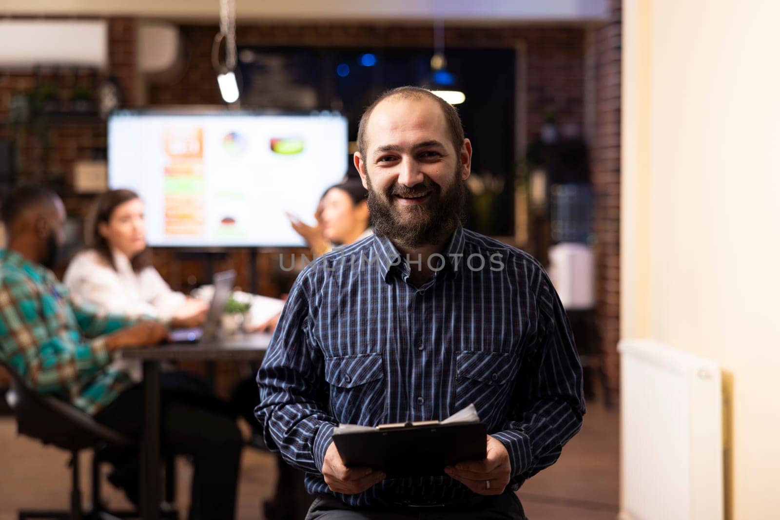 Caucasian male entrepreneur holds a clipboard in modern startup office. The night shift setting, brick wall and blurred team in background emphasize productivity and teamwork.