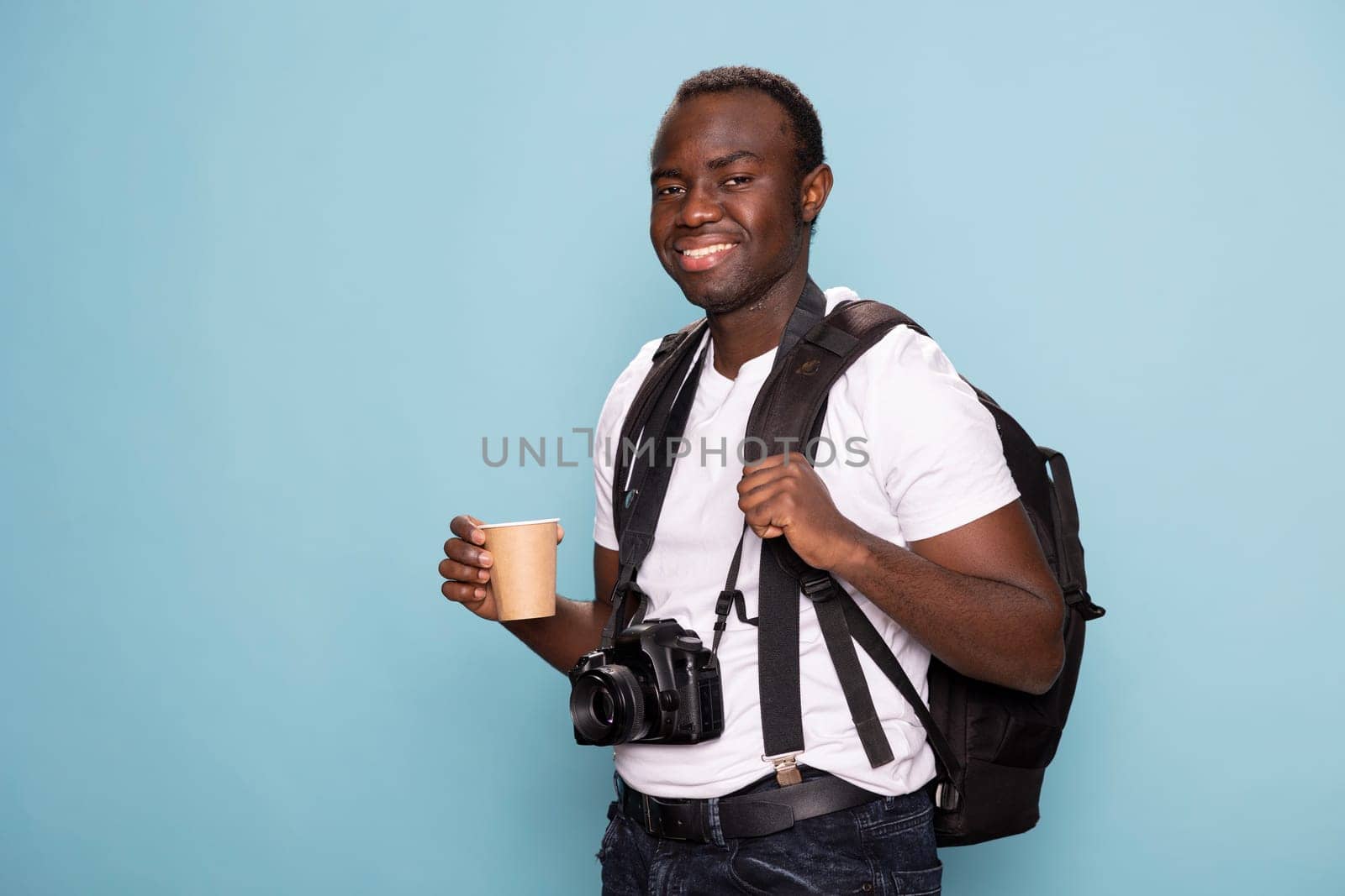 Smiling male model with a backpack and disposable cup looking directly at the camera, exuding confidence and readiness for a new creative project. Black photographer standing with his gear and coffee.