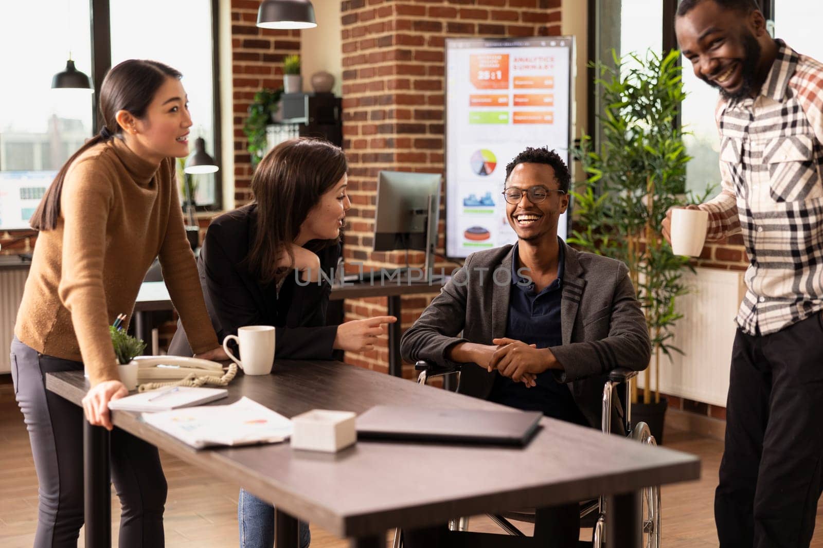 Businessman with a disability happily chatting with his diverse colleagues in a modern office. Excited employees from various backgrounds celebrating a company milestone together at startup company.