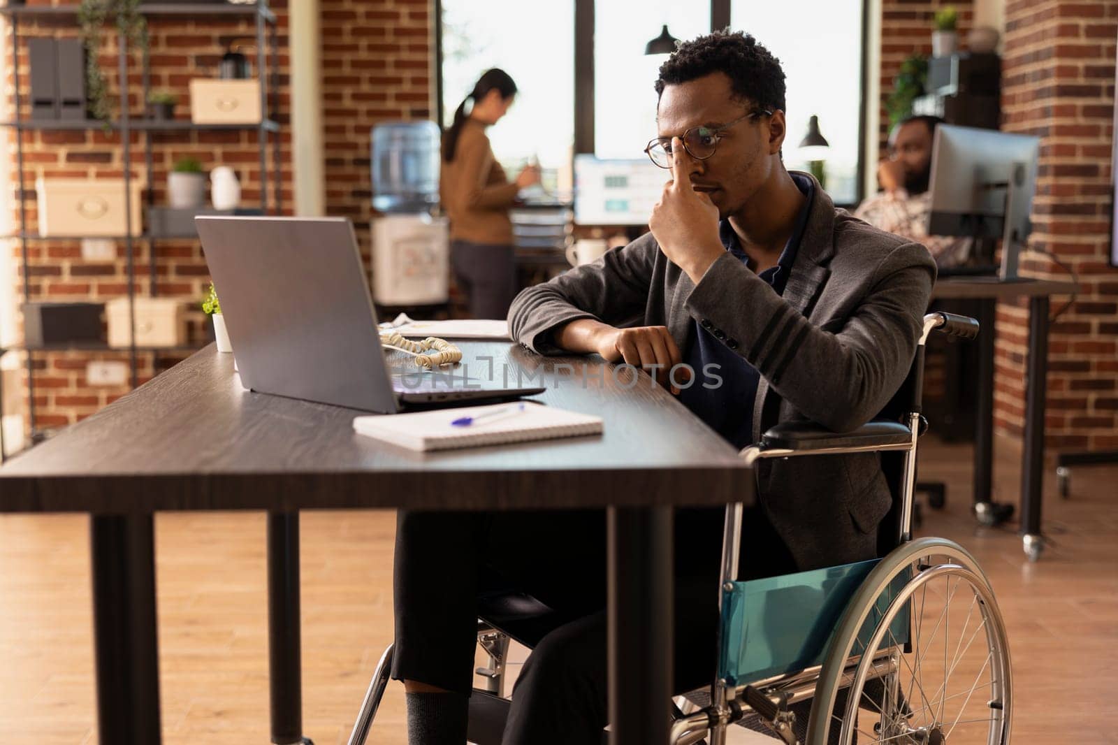 Determined employee with a physical injury, checking business analytics on his personal computer in modern workspace. Manager in wheelchair adjusting his glasses as he reviews data on his laptop.