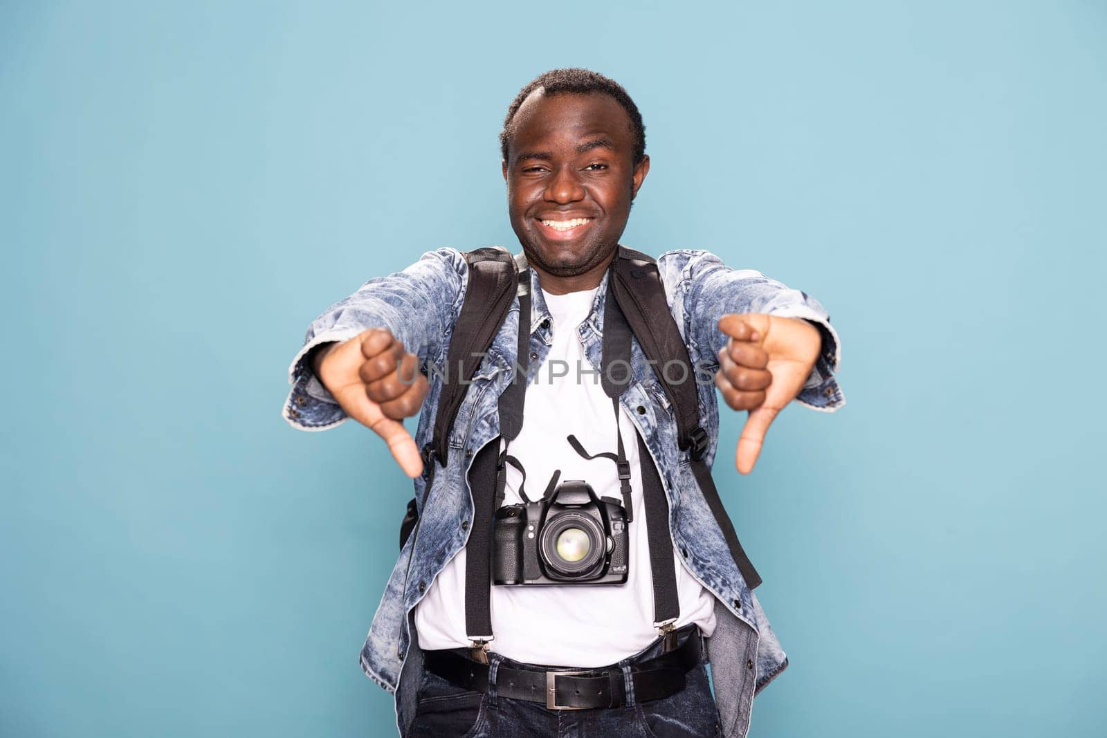 African American man in casual clothing, cheerfully giving two thumbs down while standing against isolated background. Smiling black male photographer with dslr camera, expresses disapproval in studio.