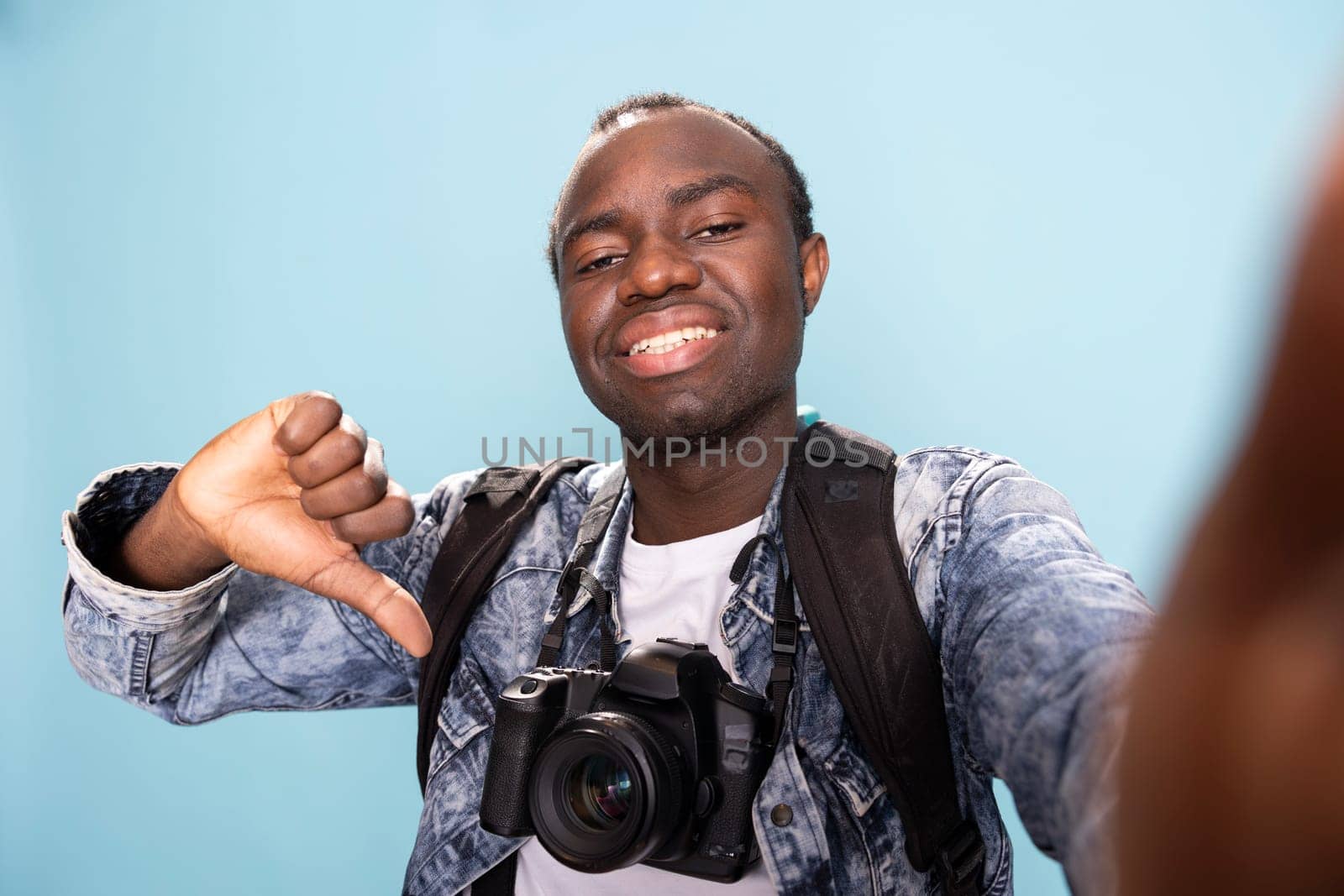 Happy african american photographer with a professional camera around his neck, giving thumbs down in studio. POV of a black male vlogger recording himself while making a playful disagreeing gesture.