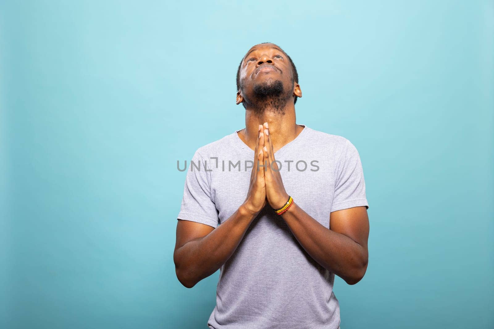 African American man with palms together, looking up and praying for good news. Black male person with hands clasped, sending a silent prayer, seeking a major achievement against isolated background.