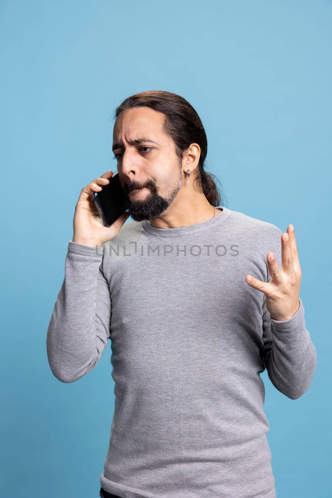 Furious young man getting mad and upset during a phone call with a friend, yelling and feeling angry against blue background. Male adult being irritated and aggressive with his words.