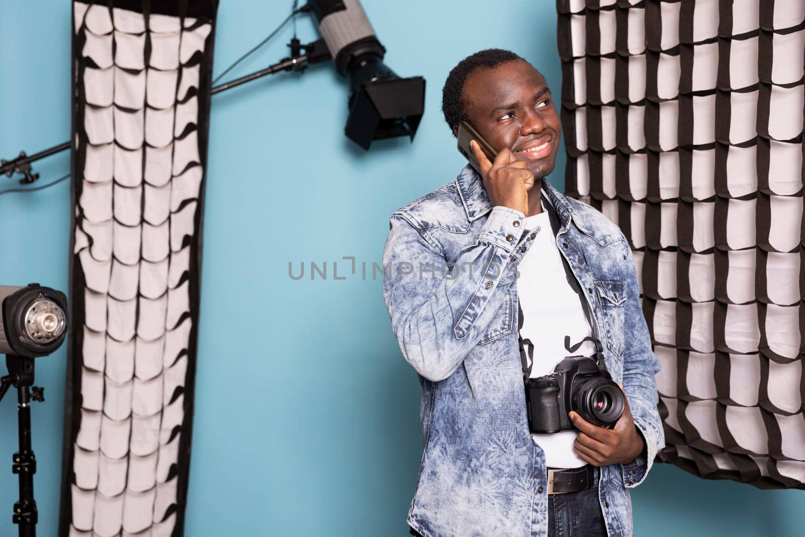 African american man grins widely while speaking on his mobile, holding his dslr device with one hand. Joyful male photographer with a camera around his neck stands, having a phone call in studio.