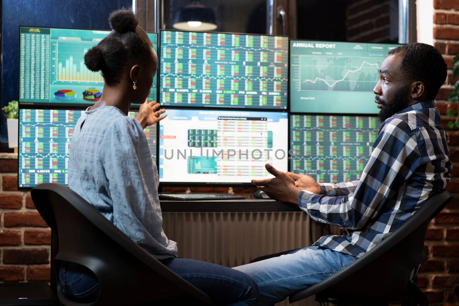 African american man and woman working late at night in office, analyzing stock market trends on multi screens. They collaborate at the desk, reviewing financial data and charts displayed on monitors.