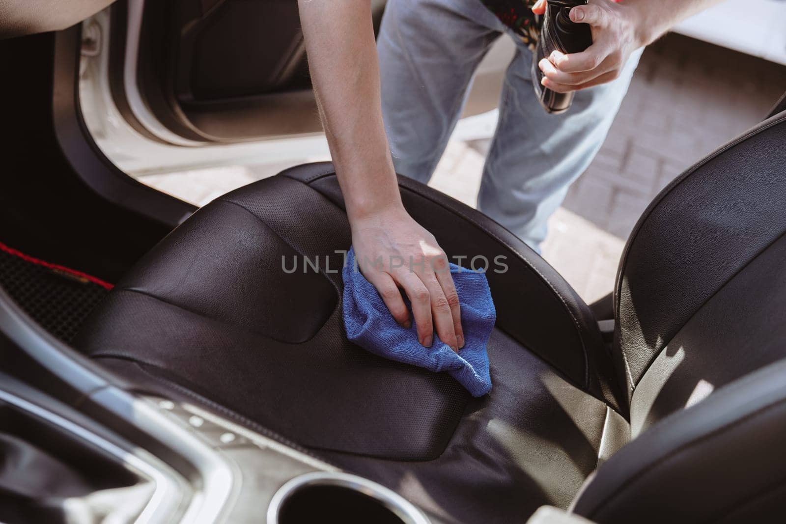 Portrait of one young Caucasian unrecognizable man who is bending over, polishing the front passenger leather seat inside the car with a blue rag on a sunny spring day at a car wash, side view close-up. Concept car wash, working people.