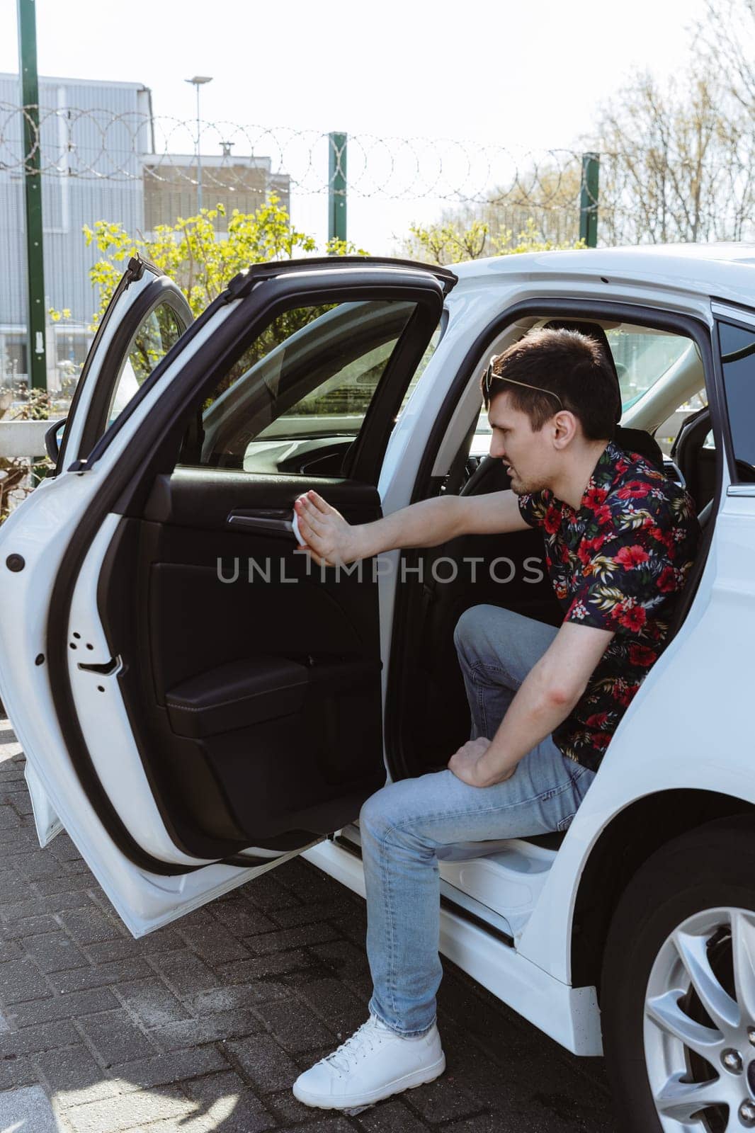 Portrait of one young Caucasian handsome man wiping leather upholstery of car passenger door with wet cloth on sunny spring day at city car wash, side view general medium plan. Concept time car wash, working people.