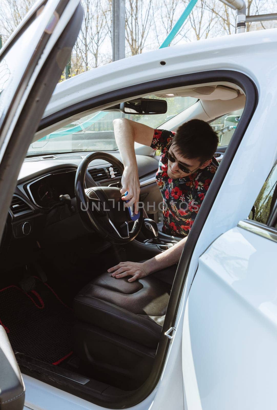 Portrait of a young handsome Caucasian man wiping the steering wheel of a car with a special sponge while sitting in the passenger seat on a sunny spring day at a city car wash, side view, medium shot. Concept: time to wash a car, working people.