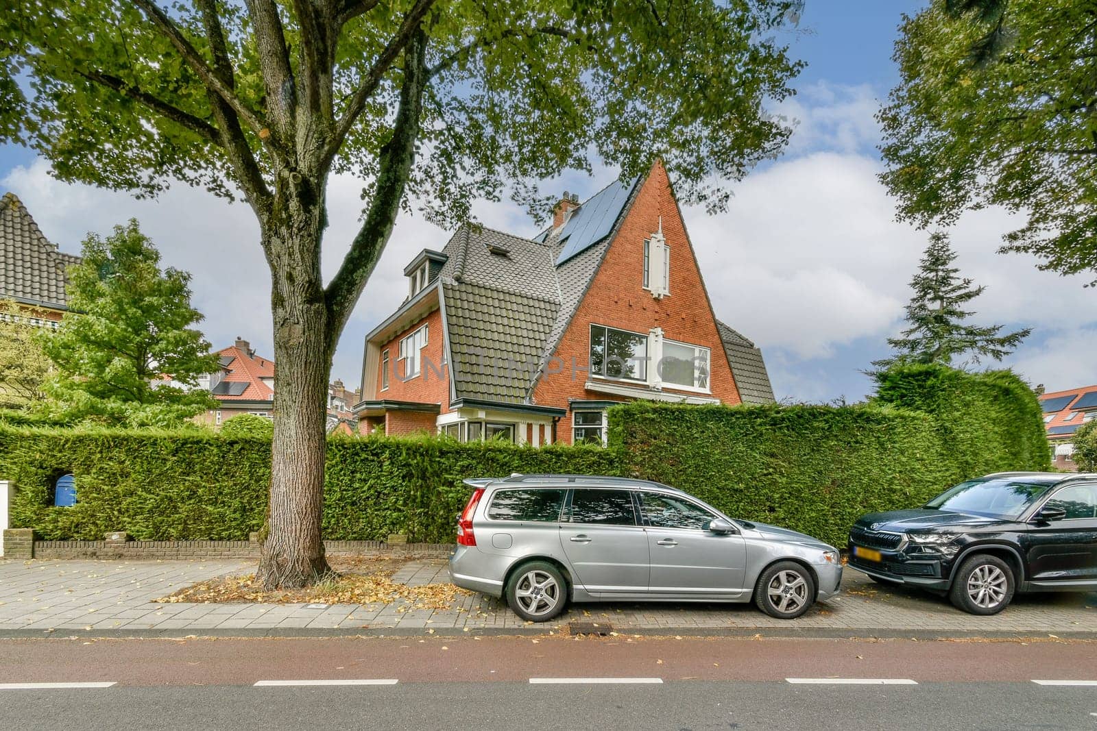 A picturesque view of a modern suburban house surrounded by lush trees and shrubs, complemented by parked cars along the street.