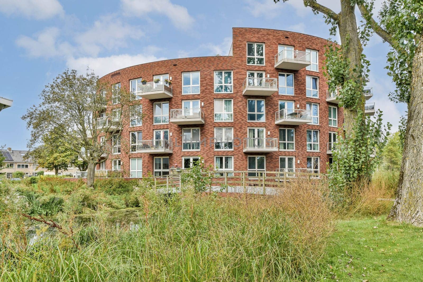 A contemporary residential building featuring large windows and balconies, harmoniously integrated with natural greenery and a serene water feature nearby.
