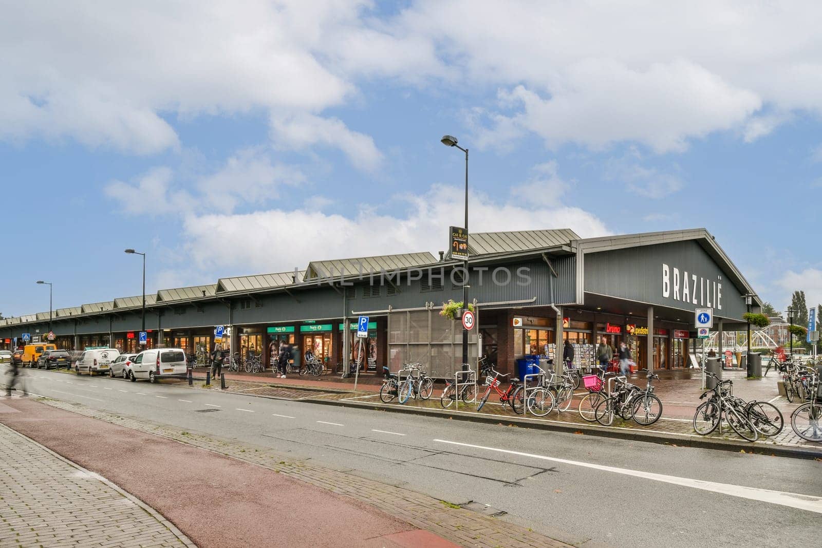 A bustling modern shopping area featuring a variety of stores, outdoor seating, and parked bicycles along a well-paved street under a clear blue sky.