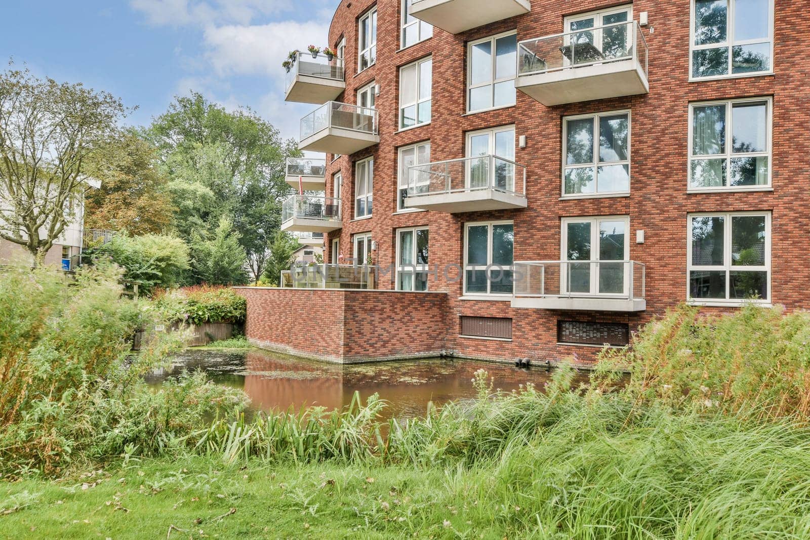 A contemporary brick apartment building features large windows and balconies, surrounded by lush greenery and a peaceful waterway for a serene atmosphere.