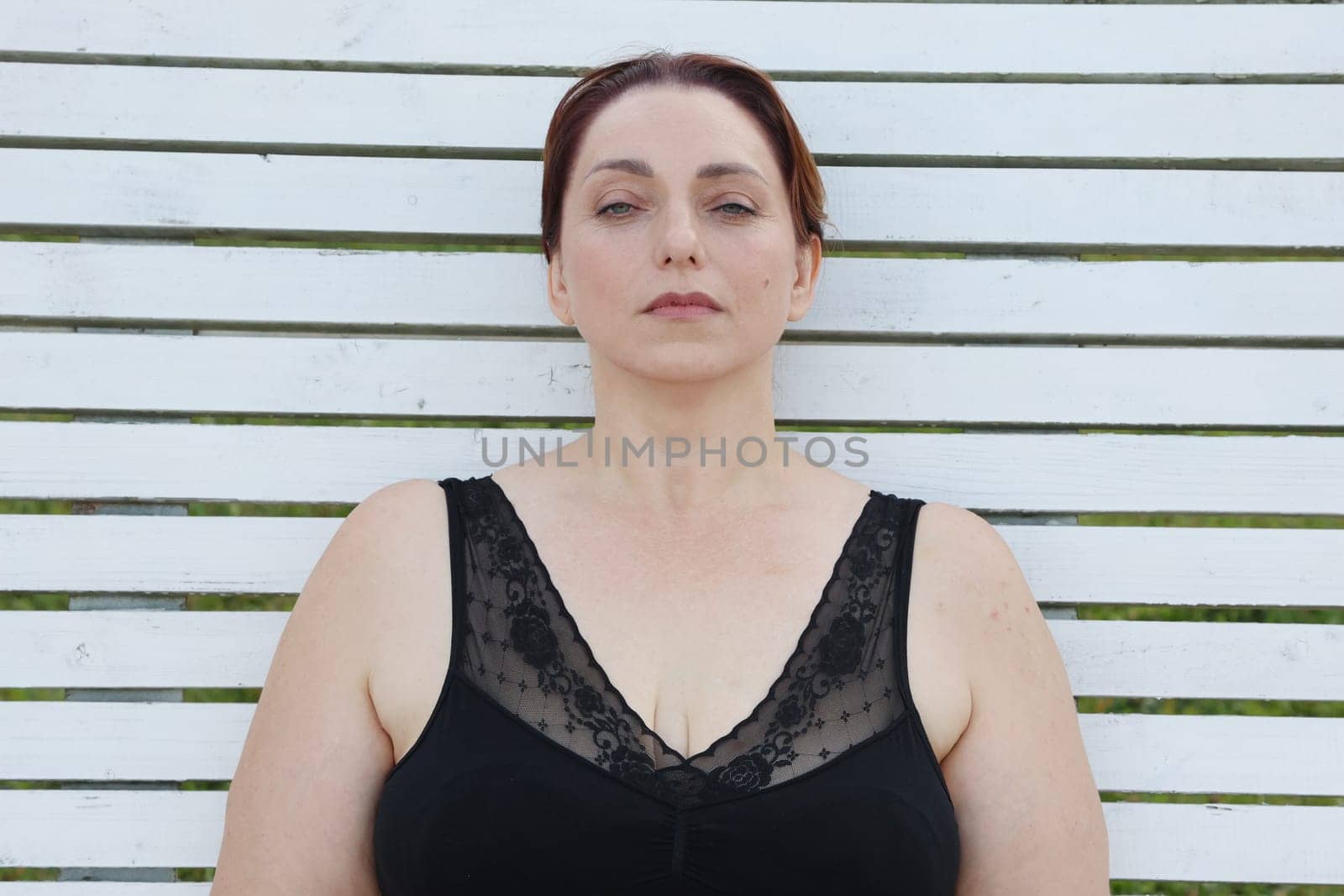 A woman in a black bra sits against a white plank wall, looking at the camera.