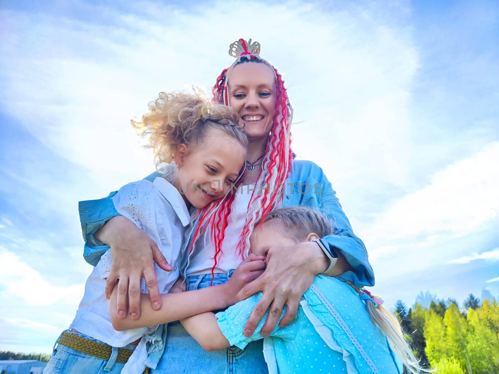 A mother joyfully holds her two daughters close, sharing smiles and laughter on a warm, sunny day outdoors.