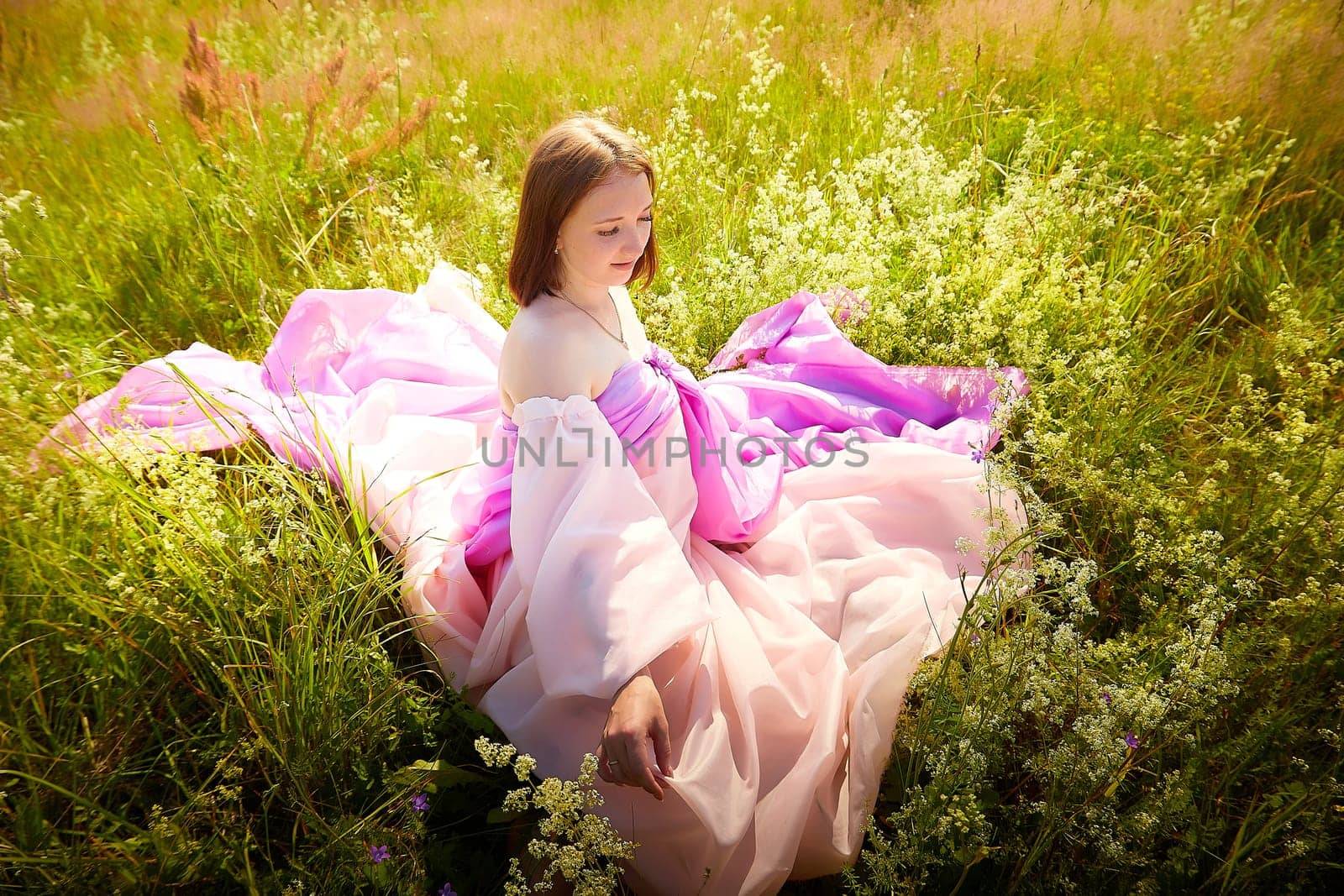 A girl with shoulder-length hair relaxes in a vibrant pink dress amidst lush green grass and blooming wildflowers under a clear blue sky.
