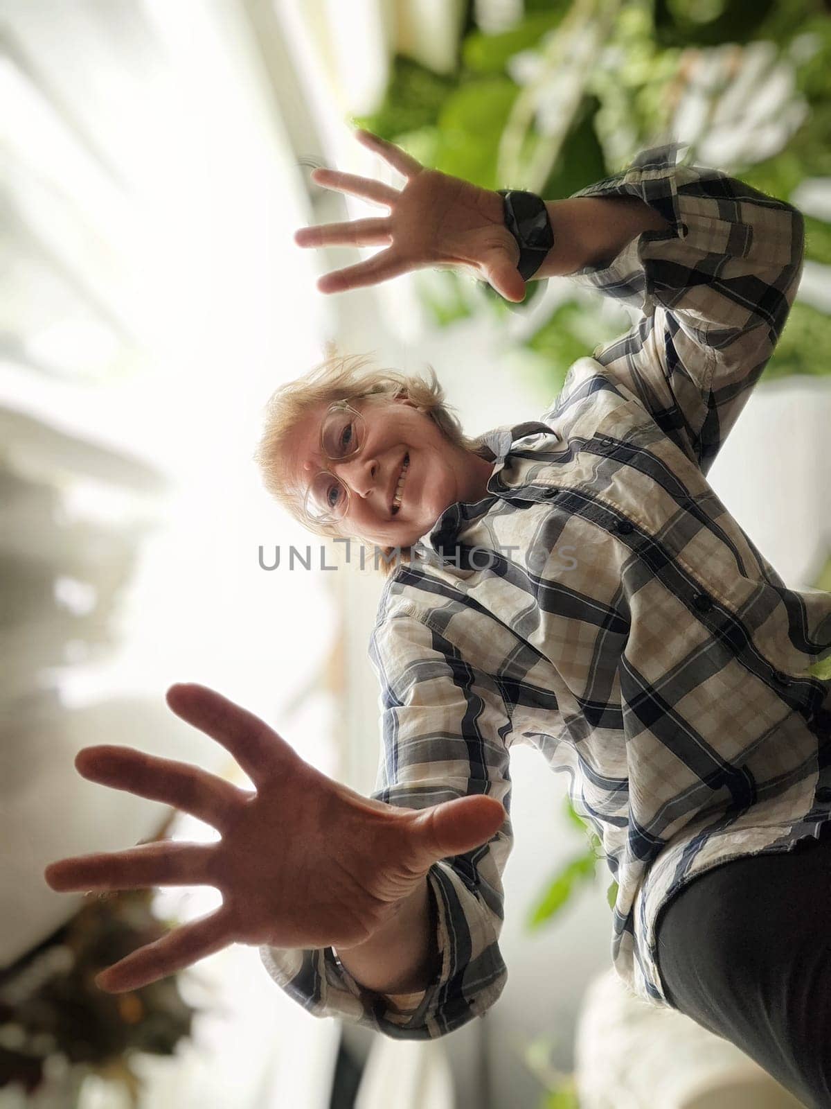 Female blogger captures a joyful selfie with outstretched hands at home, set against vibrant plants and natural light.