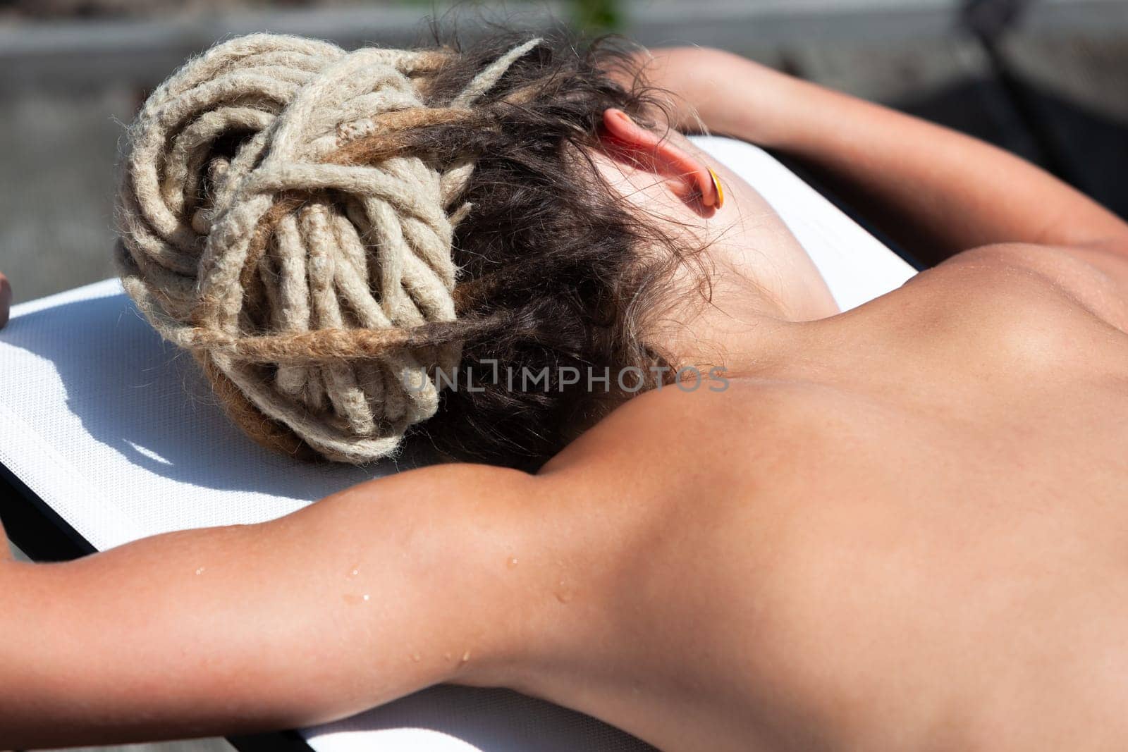 A nude woman with dreadlocks reclines on a lounge chair, enjoying the sun and a moment of relaxation outdoors.