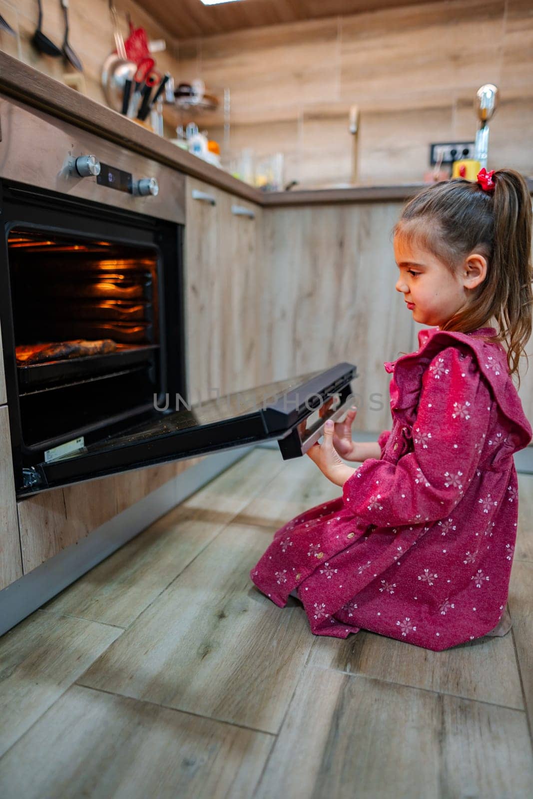 A young girl with long hair, dressed in a vibrant pink dress, kneels in front of a modern oven, eagerly watching as the door opens.