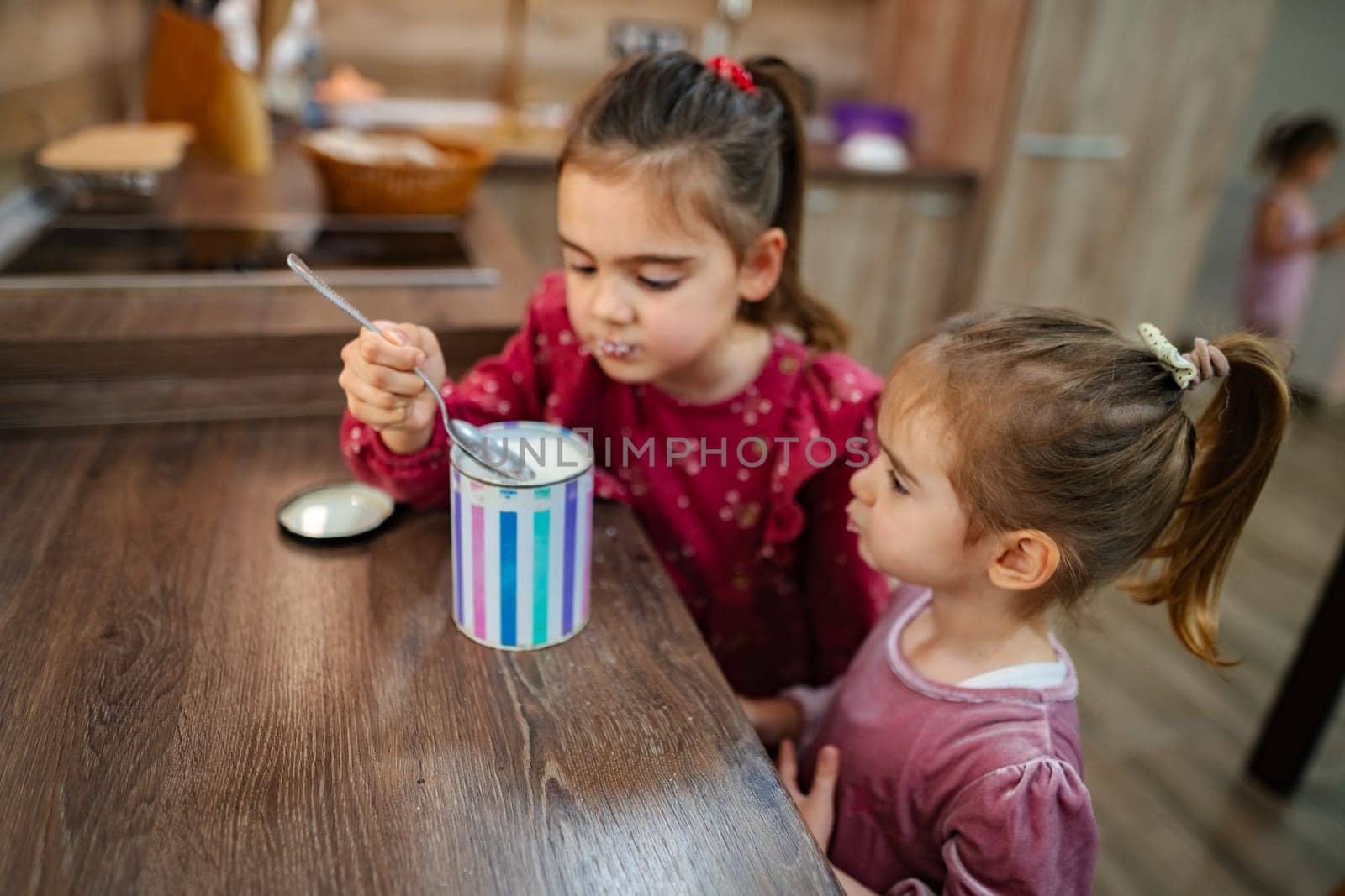 In a warm and inviting kitchen, two young girls share a delightful experience as they savor ice cream from a colorful container on a wooden table, filling their time with laughter and joy.