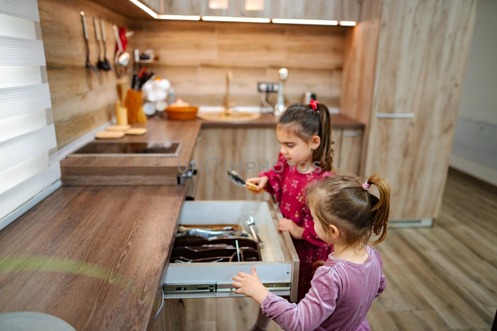 Two young sisters share a delightful moment in a modern kitchen, exploring a drawer full of utensils. Their curiosity leads to giggles as they play together, fostering creativity and bonding.