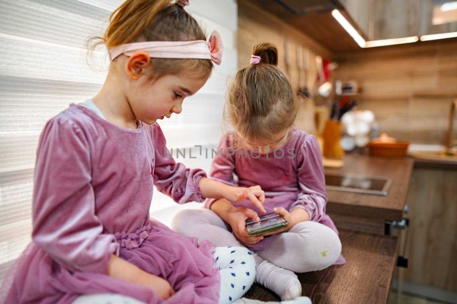 In a warm, inviting kitchen, two girls in matching dresses engage with a smartphone, sharing laughter and excitement. Natural light illuminates their joyful faces as they bond over games.