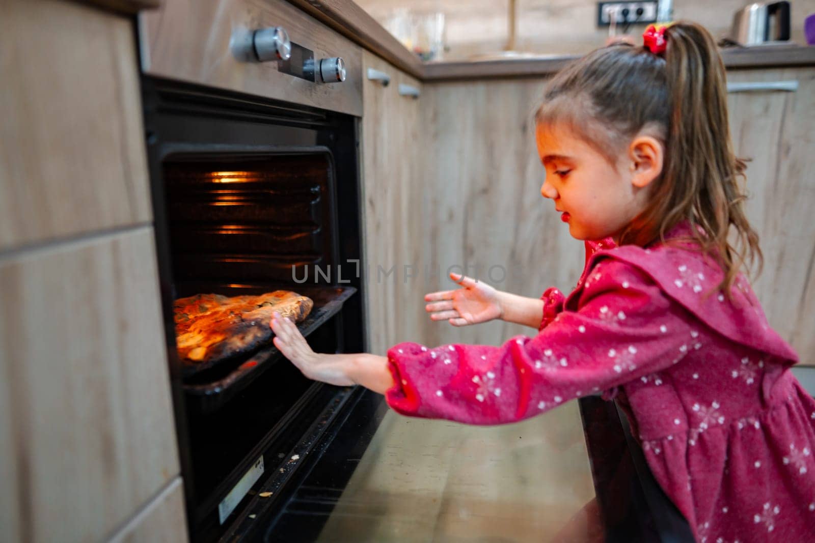 In a warm kitchen bustling with activity, a girl gleefully pulls a golden-brown pizza from the oven, her anticipation clear. The aroma fills the air as family gathers nearby.