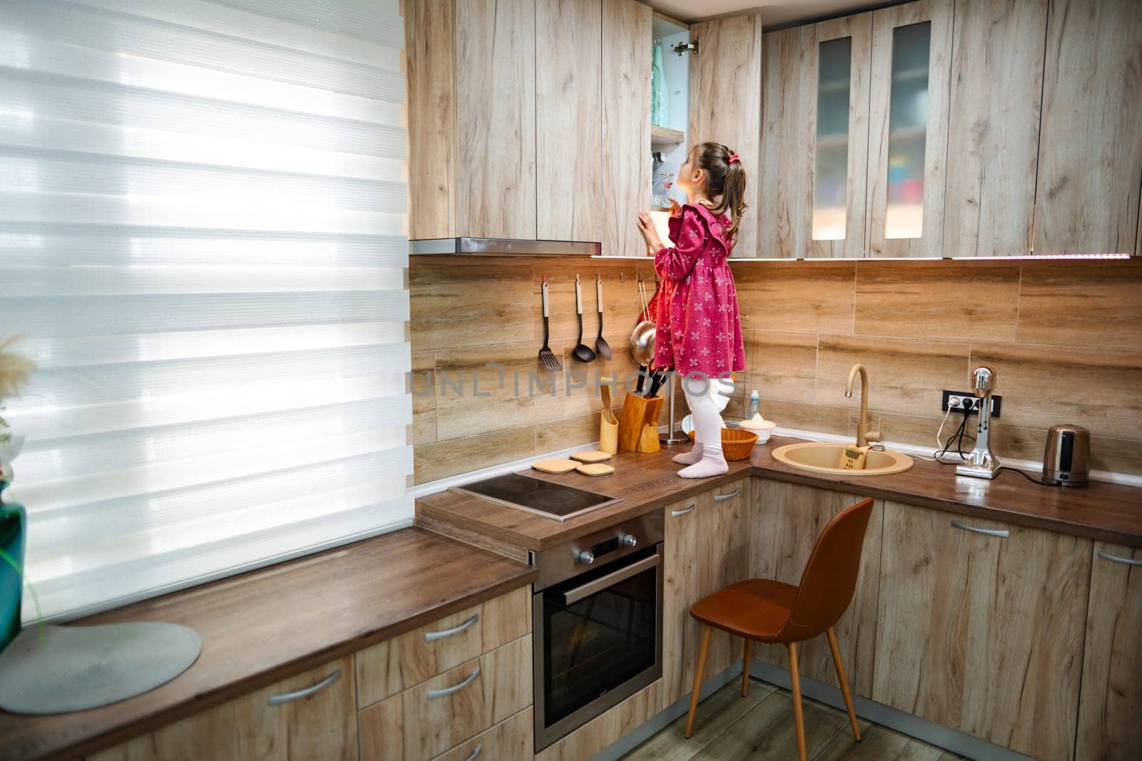 In a cozy kitchen with wooden cabinetry, a small girl in a pink dress stands on the counter, reaching up with curiosity. Sunlight streams in, adding warmth to the playful atmosphere.