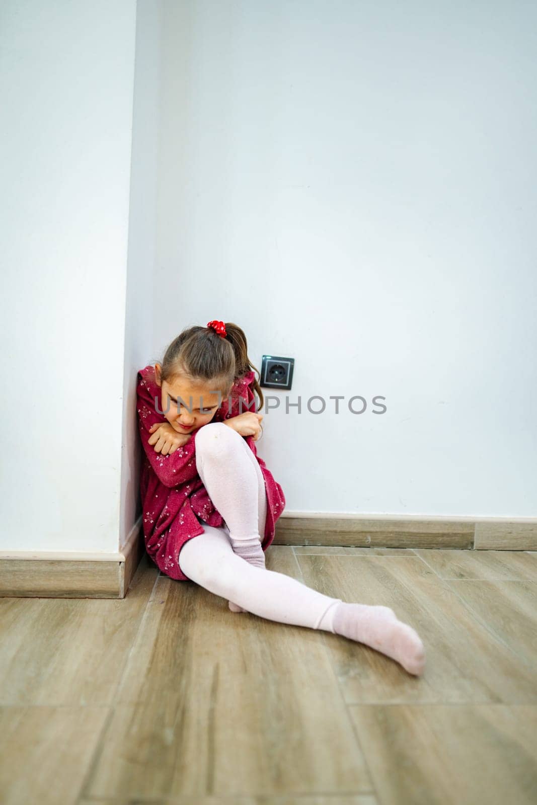 In a quiet room, a young girl huddles in the corner, wearing a playful red dress and pink tights. Her expression reveals a mix of deep thought and a desire for comfort.