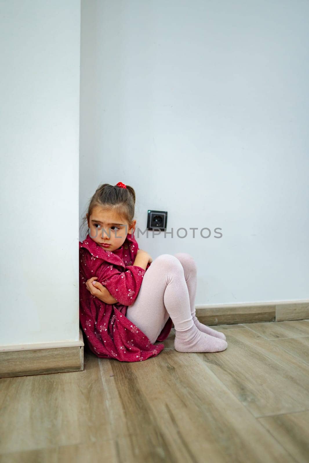 A young girl in a vibrant red dress sits curled up in a corner, her expression deep in contemplation as she gazes at the wall. The serene indoor setting enhances her solitude.
