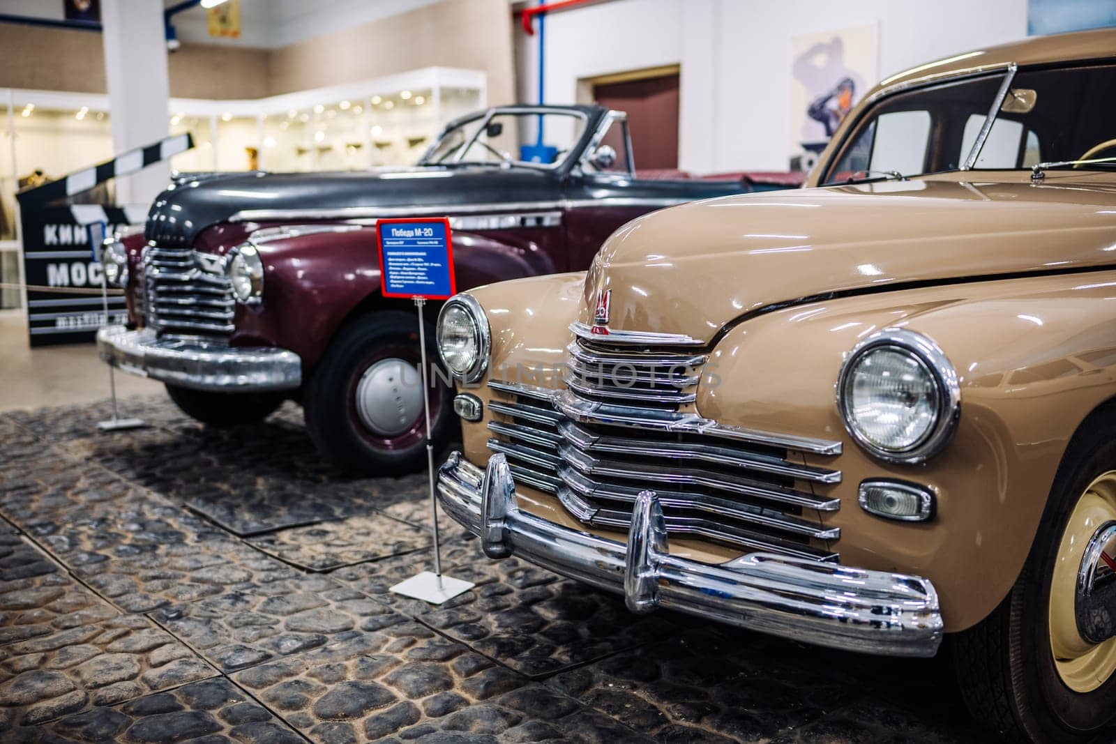 Shiny beige Pobeda car standing in front of a vintage maroon car in a museum or exhibition