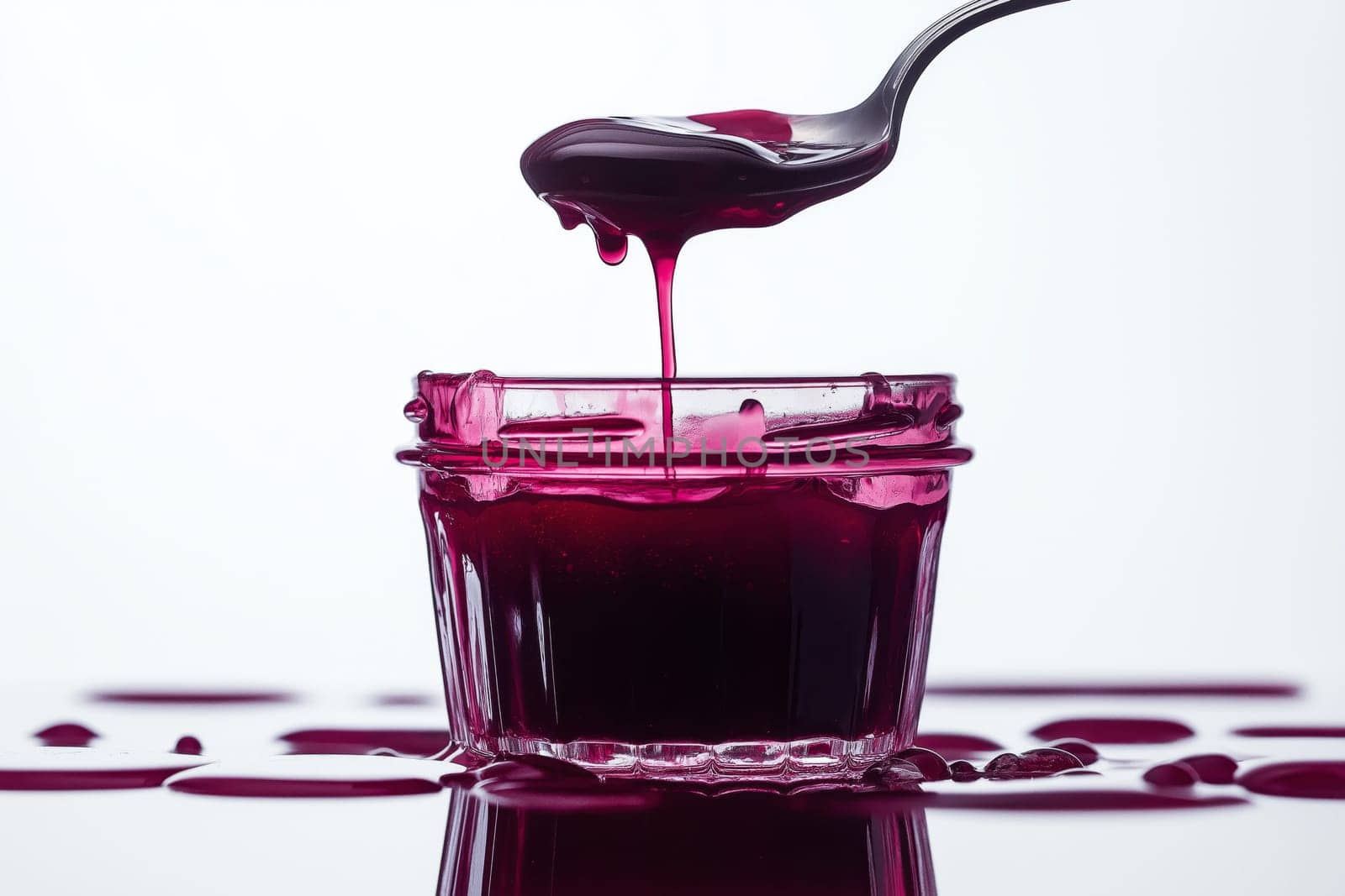 A glass jar filled with rich berry jam sits against a white background. A spoon is poised above, dripping the thick, glossy jam. Surrounding the jar are puddles of vibrant red jam.