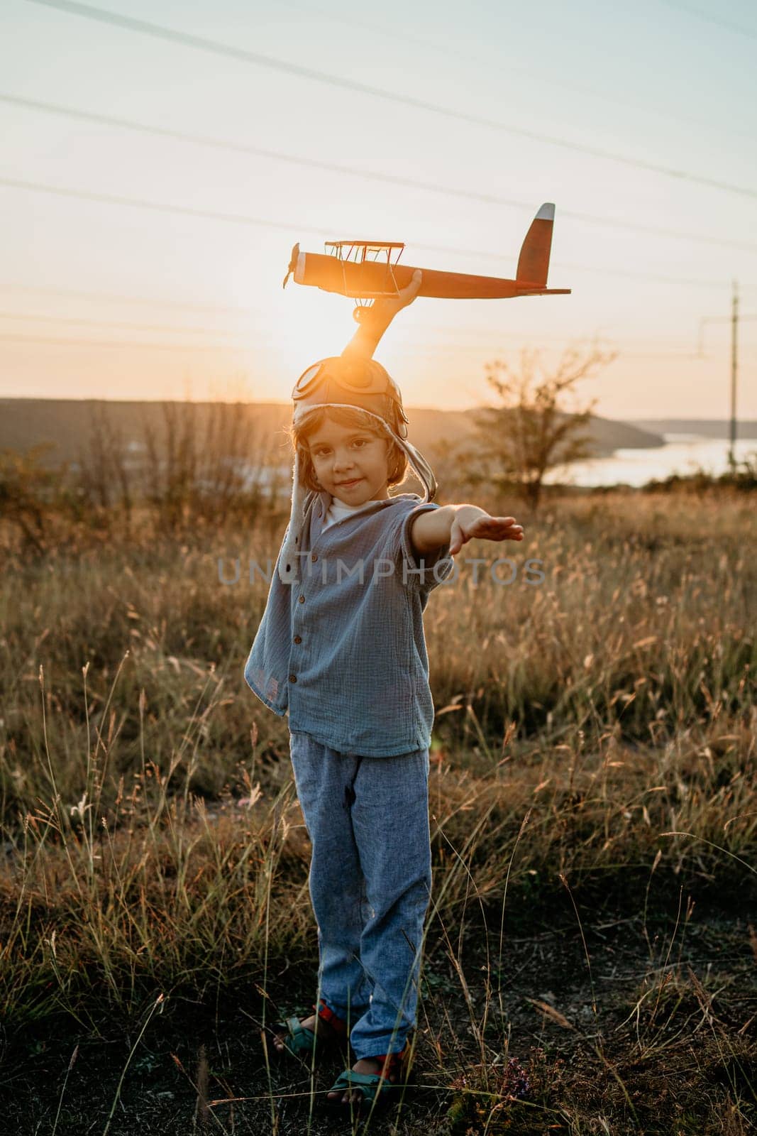 Portrait of funny little boy in aviator helmet ready to fly as airplane on nature background. Kids dream is to be pilot. Fun, creative dream, fantasy