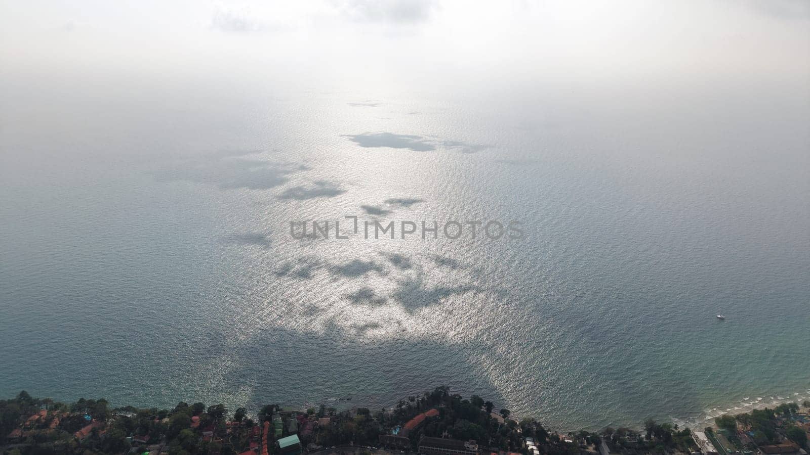 Aerial view of sun reflecting on calm ocean water, creating a mesmerizing pattern of light and shadow, with a coastal village visible at the bottom and a layer of haze obscuring the horizon