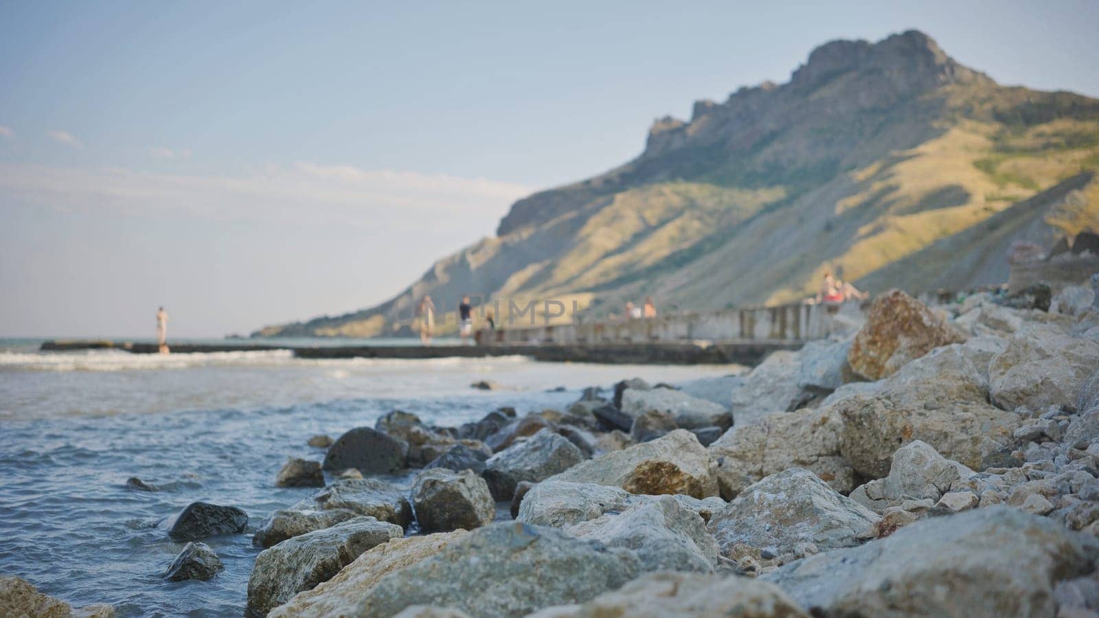Rocky beach with blurred people strolling on a pier and mountains in the background in Crimea